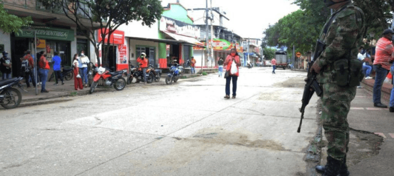 Hombre con uniforme militar vigilando en una calle concurrida del Huila, con varias personas y motos en el fondo, en medio de un contexto de inseguridad y violencia.